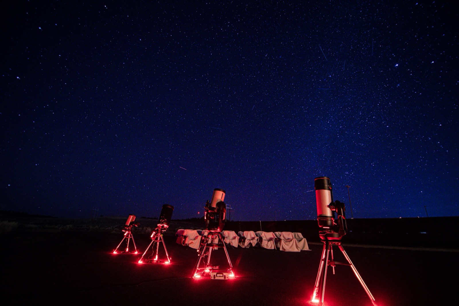 Four telescopes set up under red working lights beneath a deep blue, star-filled sky near Bryce Canyon